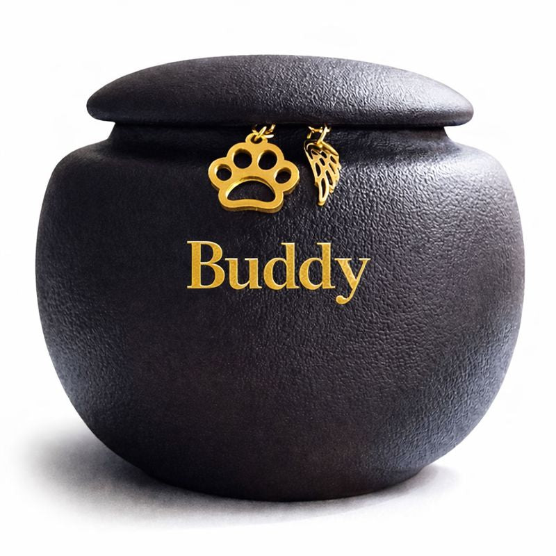 Zwarte, matte dierenurn met naam, met ronde vorm en subtiele structuur, afgewerkt met een goudkleurige pootafdruk en engelenvleugel