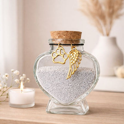Glazen hartvormige dierenurn met lichtgrijze inhoud, kurken stop en goudkleurig pootje en vleugelhanger, geplaatst op een houten dressoir met een brandend kaarsje en een rustige, minimalistische achtergrond