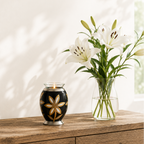 De laatste aai zwarte aluminium mini urn met gouden en zilveren lelie motief en glazen waxinelichthouder op houten tafel