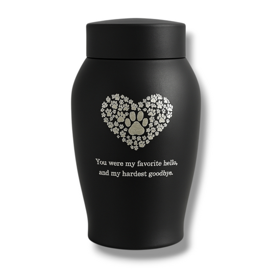 Zwarte matte urn met hart van pootafdrukken en troostende tekst, gefotografeerd tegen een lichte achtergrond.

